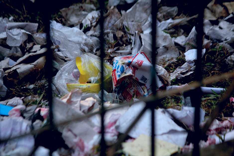 A close-up view of a pile of litter and waste materials behind a black metal fence. The waste includes various crumpled plastic bags, torn paper packaging, and discarded food wrappers, some with visible printed text and bright colours such as yellow, red, and white. The trash is scattered on the ground amid dead leaves and organic debris, with the fence posts and bars partially obscuring the view. The environment appears outdoor, on or near a pathway or garden area, suggesting a location where illegal dumping or unmanaged rubbish collection might occur. Waste Removal Swiss Cottage offers services that address this type of rubbish accumulation, supporting private waste clearance solutions outside of council-managed rubbish collection, as evidenced by the type and unmanaged state of the debris visible in the image.