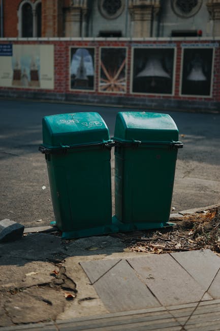A close-up view of a pile of litter and waste materials behind a black metal fence. The waste includes various crumpled plastic bags, torn paper packaging, and discarded food wrappers, some with visible printed text and bright colours such as yellow, red, and white. The trash is scattered on the ground amid dead leaves and organic debris, with the fence posts and bars partially obscuring the view. The environment appears outdoor, on or near a pathway or garden area, suggesting a location where illegal dumping or unmanaged rubbish collection might occur. Waste Removal Swiss Cottage offers services that address this type of rubbish accumulation, supporting private waste clearance solutions outside of council-managed rubbish collection, as evidenced by the type and unmanaged state of the debris visible in the image.