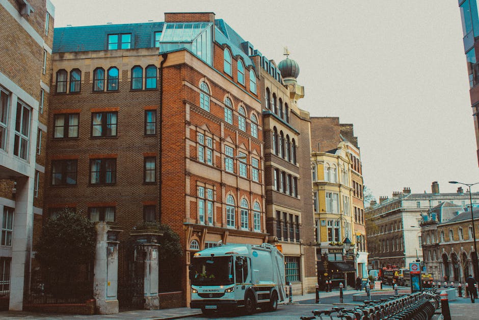 The image depicts a narrow city street lined with multi-story buildings showcasing a mix of architectural styles, including brick facades, arched windows, and decorative cornices. On the left side, a building features a brown brick exterior with several rectangular windows, partially obscured by dark green trees and a stone wall with a metal gate. The central focus is a small, light blue refuse collection truck parked on the street, positioned in front of a modern glass-fronted building. The street scene includes street furniture such as bicycle racks and bollards, with a few pedestrians visible in the background, alongside additional historic and contemporary buildings. The bright, overcast sky creates diffuse lighting, highlighting the textures of the building surfaces and the smooth finish of the waste collection vehicle. The image reflects an urban environment where private waste management services, like Waste Removal Swiss Cottage, operate within a historic and modern cityscape, emphasizing the importance of independent rubbish collection and on-site clearance in such areas.